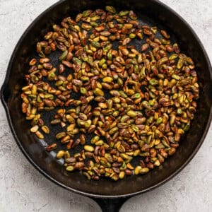 A black cast iron skillet filled with roasted pistachios sits on a light textured surface.