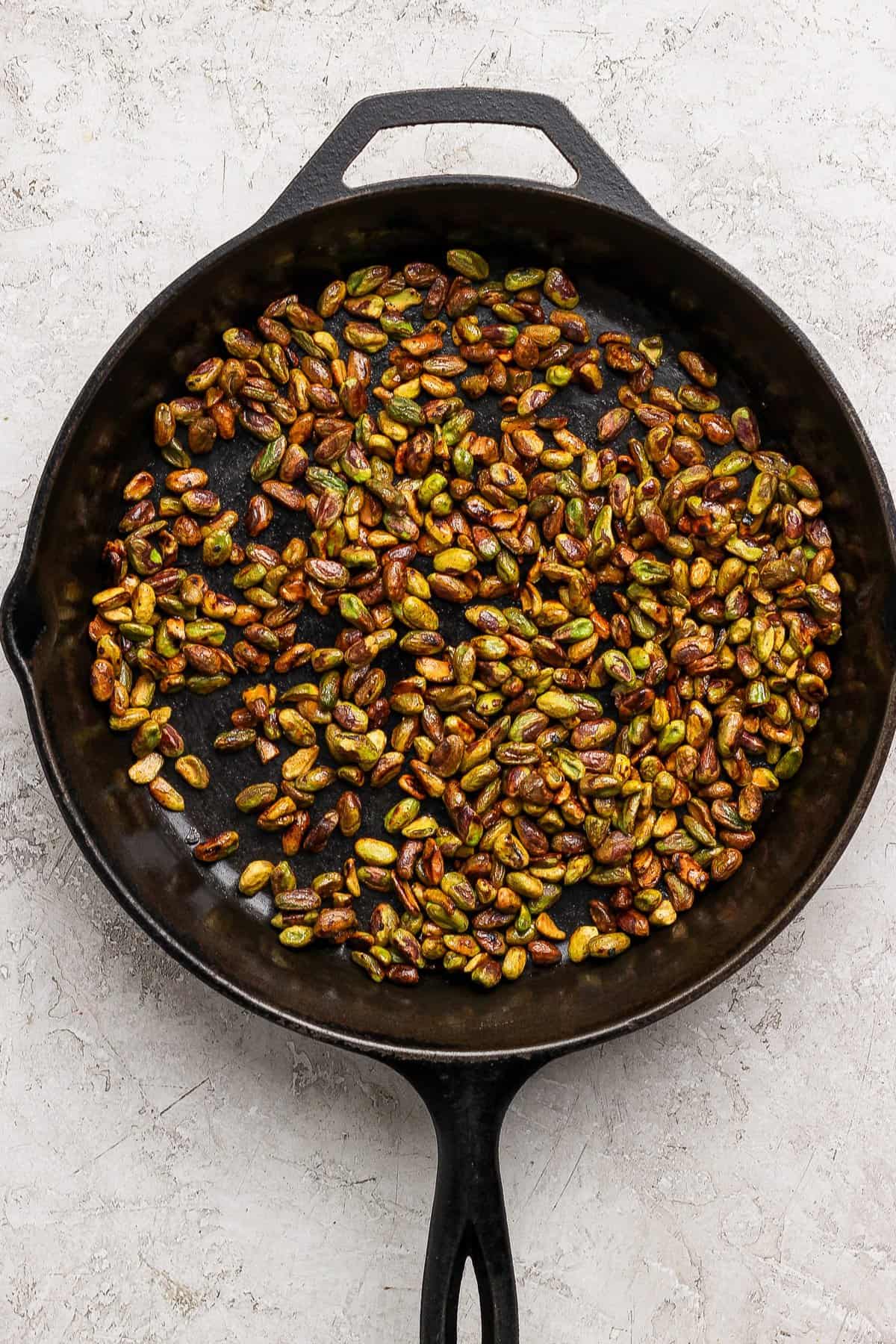 A black cast iron skillet filled with roasted pistachios sits on a light textured surface.