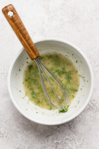 A white bowl containing a light-colored vinaigrette with herbs, being mixed with a metal whisk with a wooden handle, set on a light-textured surface.