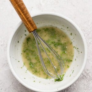 A white bowl containing a light-colored vinaigrette with herbs, being mixed with a metal whisk with a wooden handle, set on a light-textured surface.