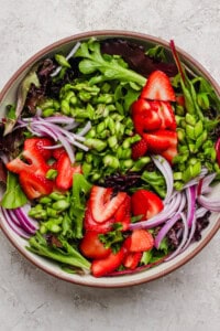 A bowl of mixed greens salad topped with sliced strawberries, red onion, and chopped green vegetables on a light textured surface.