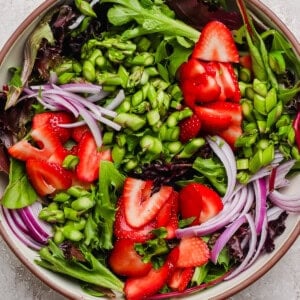 A bowl of mixed greens salad topped with sliced strawberries, red onion, and chopped green vegetables on a light textured surface.