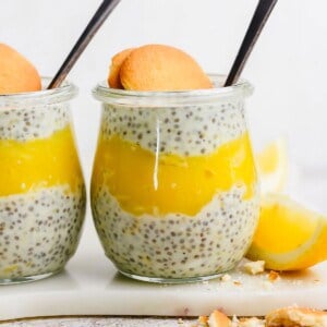 A glass jar layered with chia pudding and yellow lemon curd, topped with a round cookie and a spoon, with a lemon wedge next to the jar.
