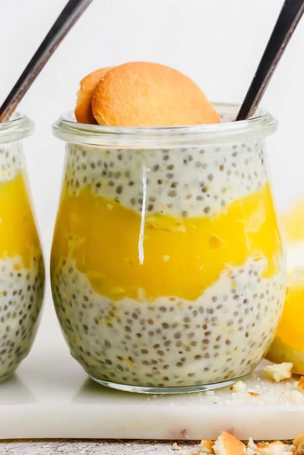 A glass jar layered with chia pudding and yellow lemon curd, topped with a round cookie and a spoon, with a lemon wedge next to the jar.