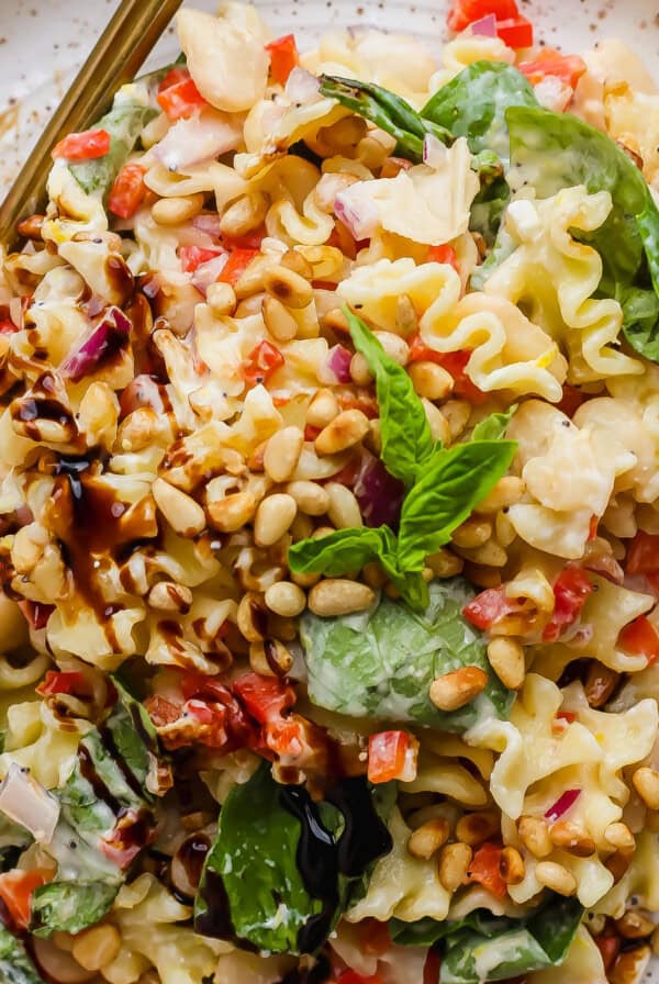 A plate of pasta salad with spinach, pine nuts, white beans, chopped red onions, red bell peppers, and a balsamic drizzle, garnished with fresh basil.