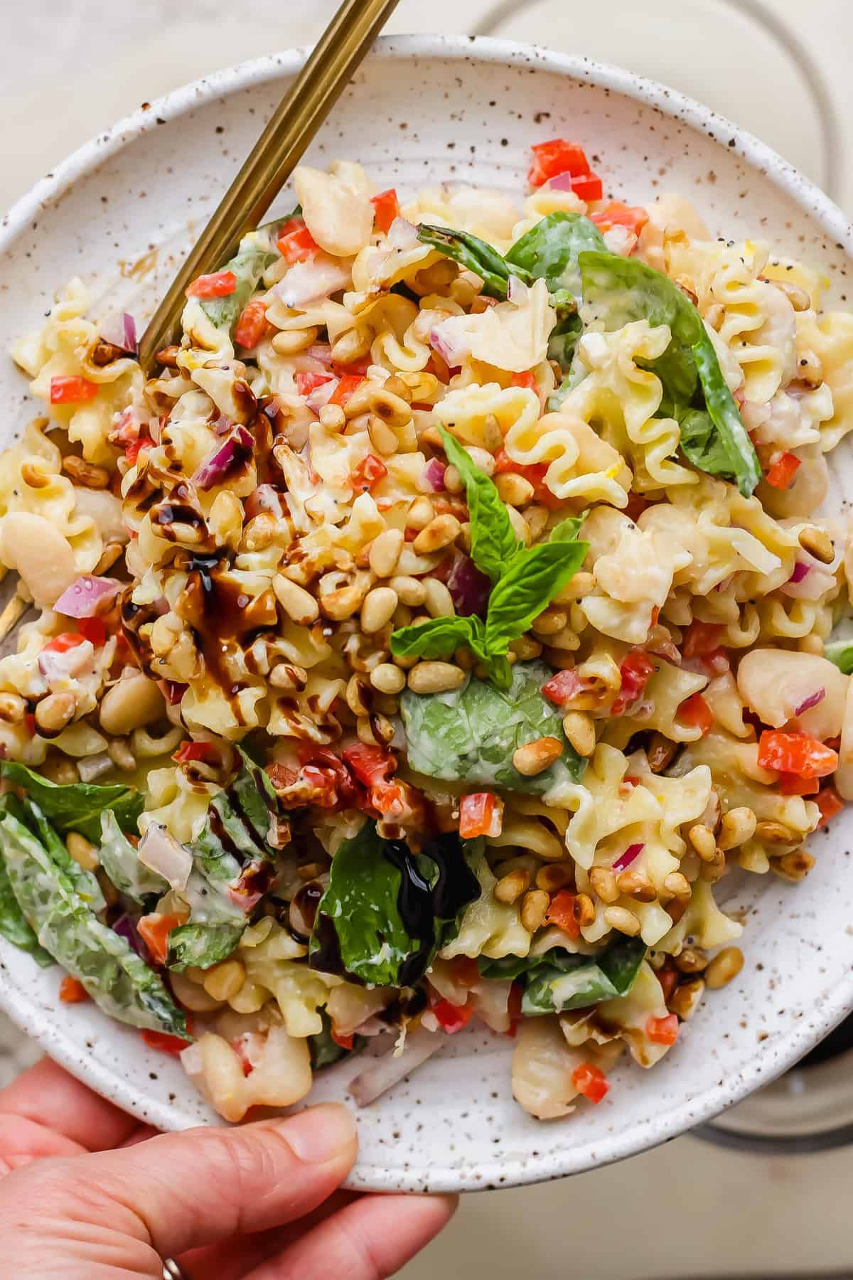 A plate of pasta salad with spinach, cannellini beans, red onion, diced red peppers, pine nuts, and a balsamic glaze, served with a fork.