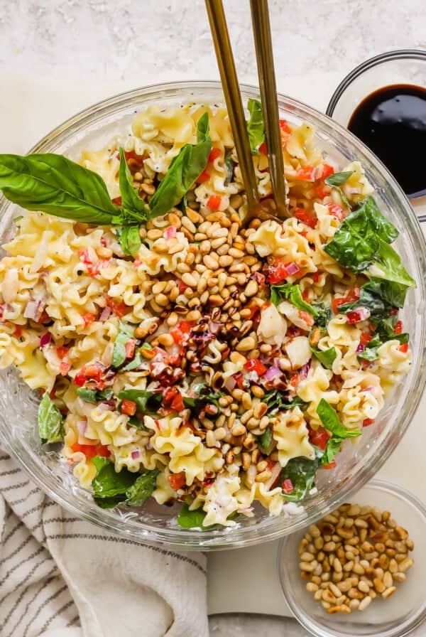 A glass bowl of pasta salad with spinach, pine nuts, red onion, and chopped vegetables, garnished with basil leaves and served with dressing on the side.