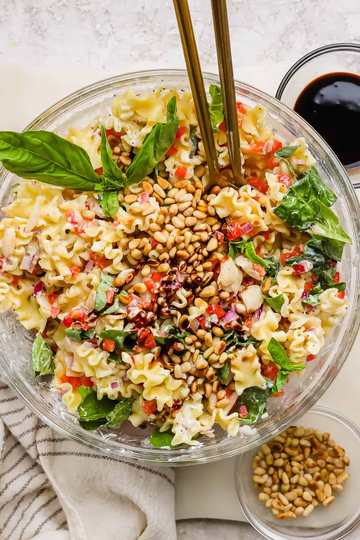 A glass bowl of pasta salad with spinach, pine nuts, red onion, and chopped vegetables, garnished with basil leaves and served with dressing on the side.