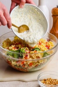 A hand pours creamy dressing from a bowl onto a pasta salad with vegetables in a clear glass mixing bowl. A bowl of pine nuts sits nearby.