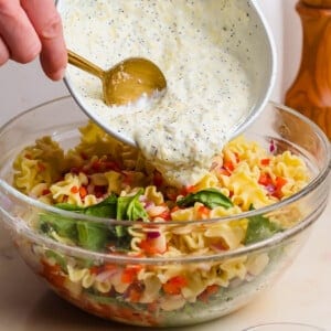 A hand pours creamy dressing from a bowl onto a pasta salad with vegetables in a clear glass mixing bowl. A bowl of pine nuts sits nearby.