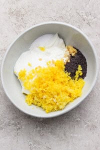 A bowl containing yogurt, lemon zest, poppy seeds, minced garlic, and salt on a light-colored surface.