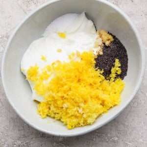 A bowl containing yogurt, lemon zest, poppy seeds, minced garlic, and salt on a light-colored surface.