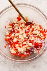 A clear bowl containing a mixture of white beans, diced red bell pepper, and chopped red onion, with a spoon for stirring.