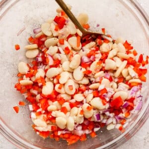 A clear bowl containing a mixture of white beans, diced red bell pepper, and chopped red onion, with a spoon for stirring.