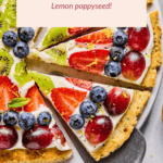 A fruit pizza topped with blueberries, strawberries, kiwi, and grapes on a lemon poppy seed crust, sliced and served on a plate with lemon slices and strawberries nearby.