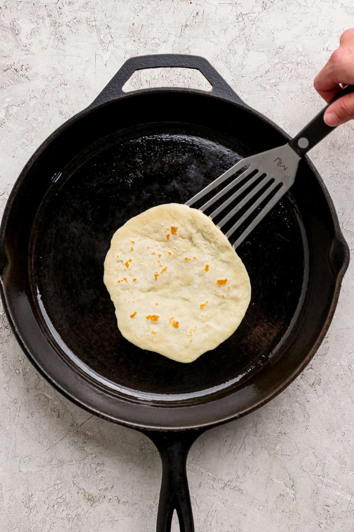 A hand uses a spatula to lift a tortilla cooking in a black cast iron skillet on a light-colored surface.