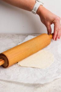 A hand holding a rolling pin rolls out dough on parchment paper on a light-colored surface.