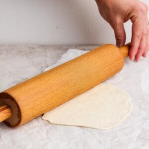 A hand holding a rolling pin rolls out dough on parchment paper on a light-colored surface.
