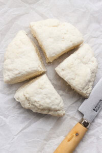 Four wedges of raw biscuit dough and a knife with a wooden handle on white parchment paper.