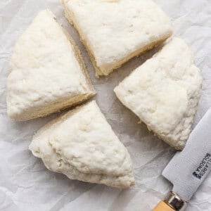 Four wedges of raw biscuit dough and a knife with a wooden handle on white parchment paper.