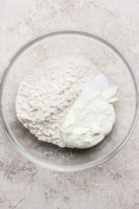 A glass bowl containing flour, white powder (possibly salt or baking powder), and a dollop of plain yogurt on a light textured surface.