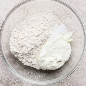 A glass bowl containing flour, white powder (possibly salt or baking powder), and a dollop of plain yogurt on a light textured surface.