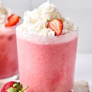 A glass of pink strawberry smoothie topped with whipped cream and a sliced strawberry, with whole strawberries nearby.