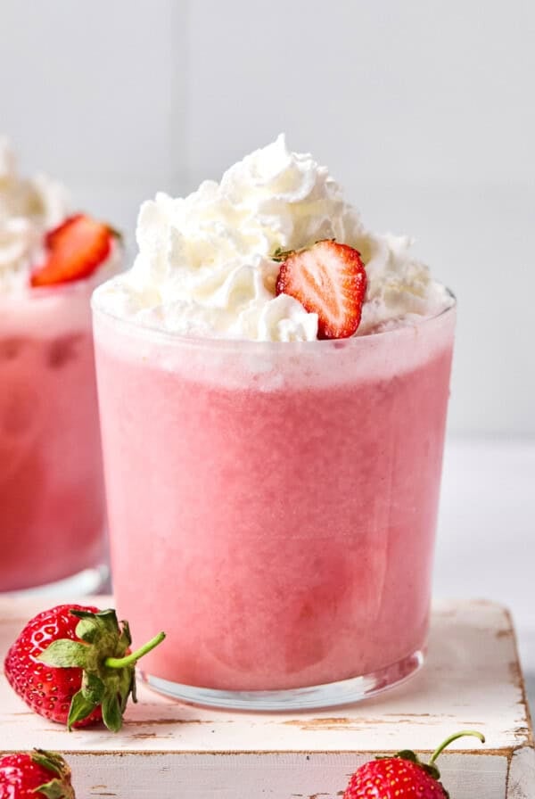 A glass of pink strawberry smoothie topped with whipped cream and a sliced strawberry, with whole strawberries nearby.