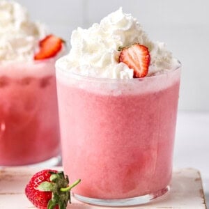 A glass of pink strawberry milkshake topped with whipped cream and a sliced strawberry, with another drink and a whole strawberry nearby.