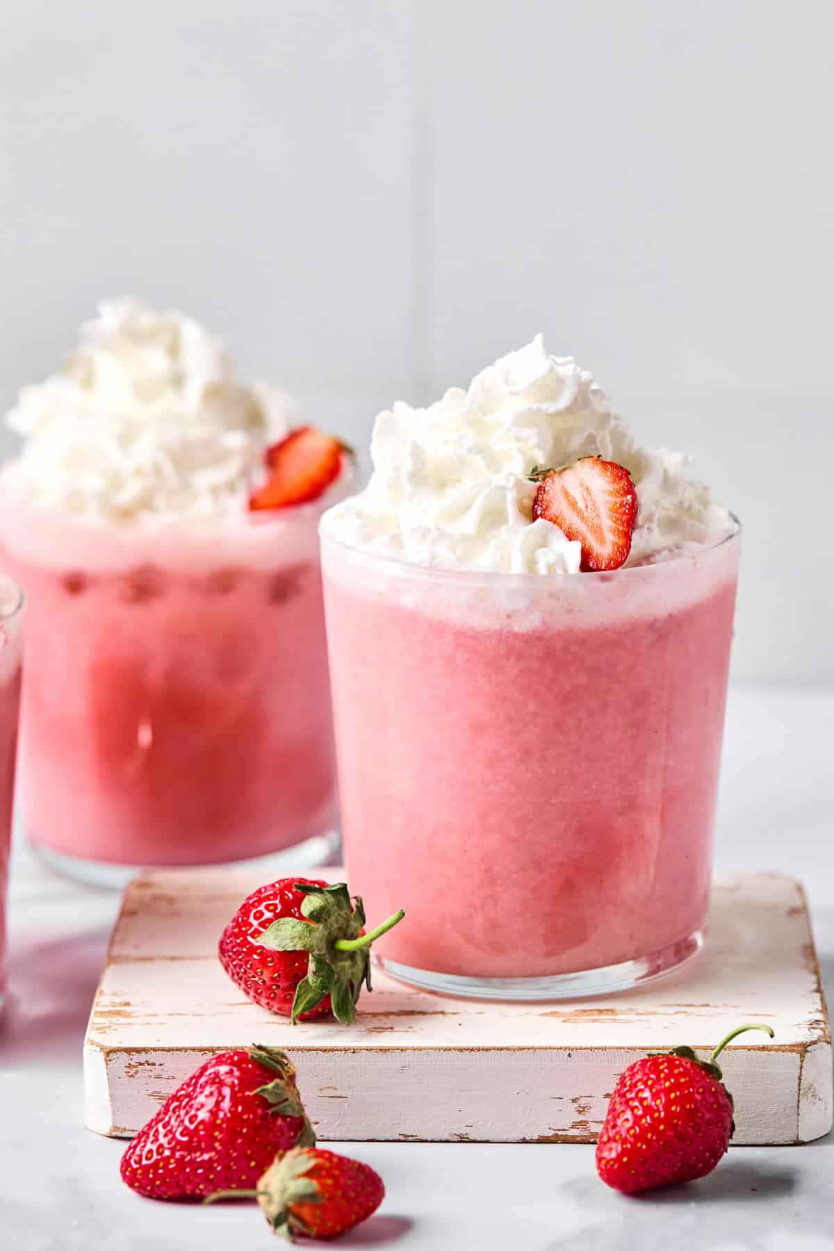 A glass of pink strawberry smoothie topped with whipped cream and a strawberry slice, with fresh strawberries placed beside the glass.