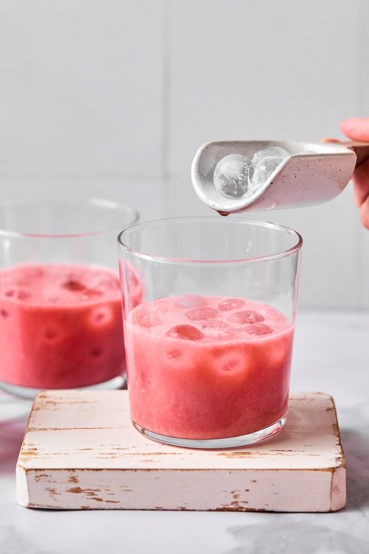 A hand adds ice cubes from a small white scoop into a glass of pink iced beverage, with another glass of the same drink in the background.