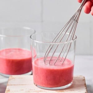A hand uses a metal whisk to mix a pink liquid in a glass, with another glass containing the same liquid in the background on a white surface.