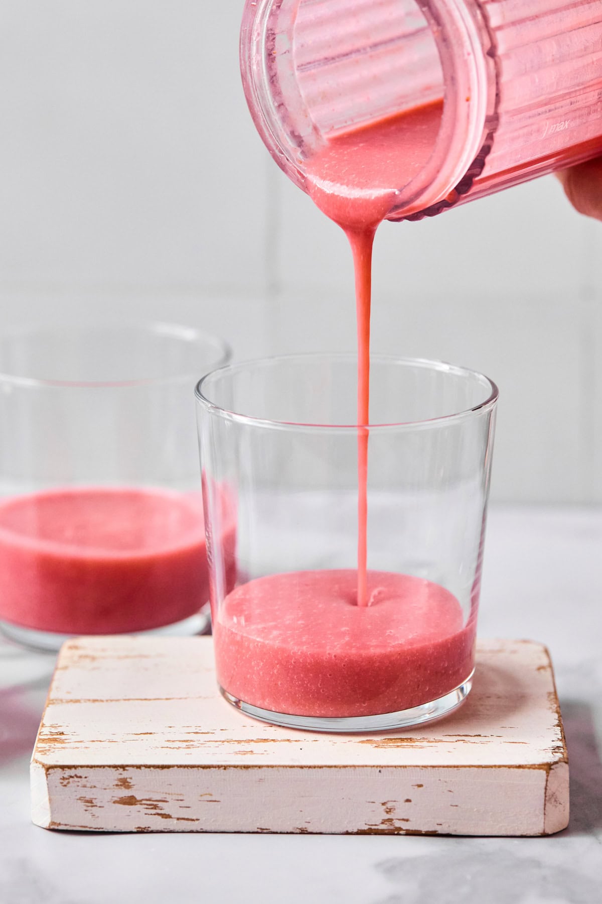 Pink smoothie being poured from a blender into a glass, with another glass of smoothie nearby on a small white wooden board.
