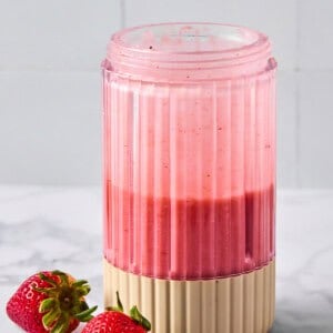 A pink smoothie in a ribbed glass sits on a marble surface next to two fresh strawberries.