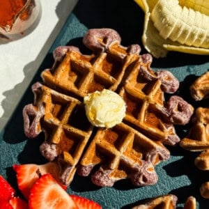A waffle topped with a dollop of butter is surrounded by sliced bananas and strawberries on a dark surface.