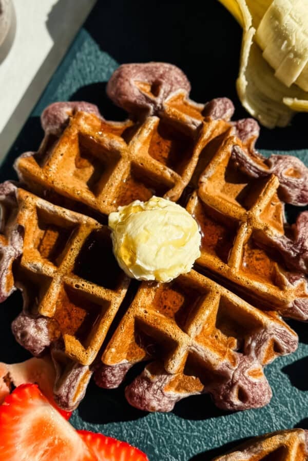 A waffle topped with a dollop of butter is surrounded by sliced bananas and strawberries on a dark surface.