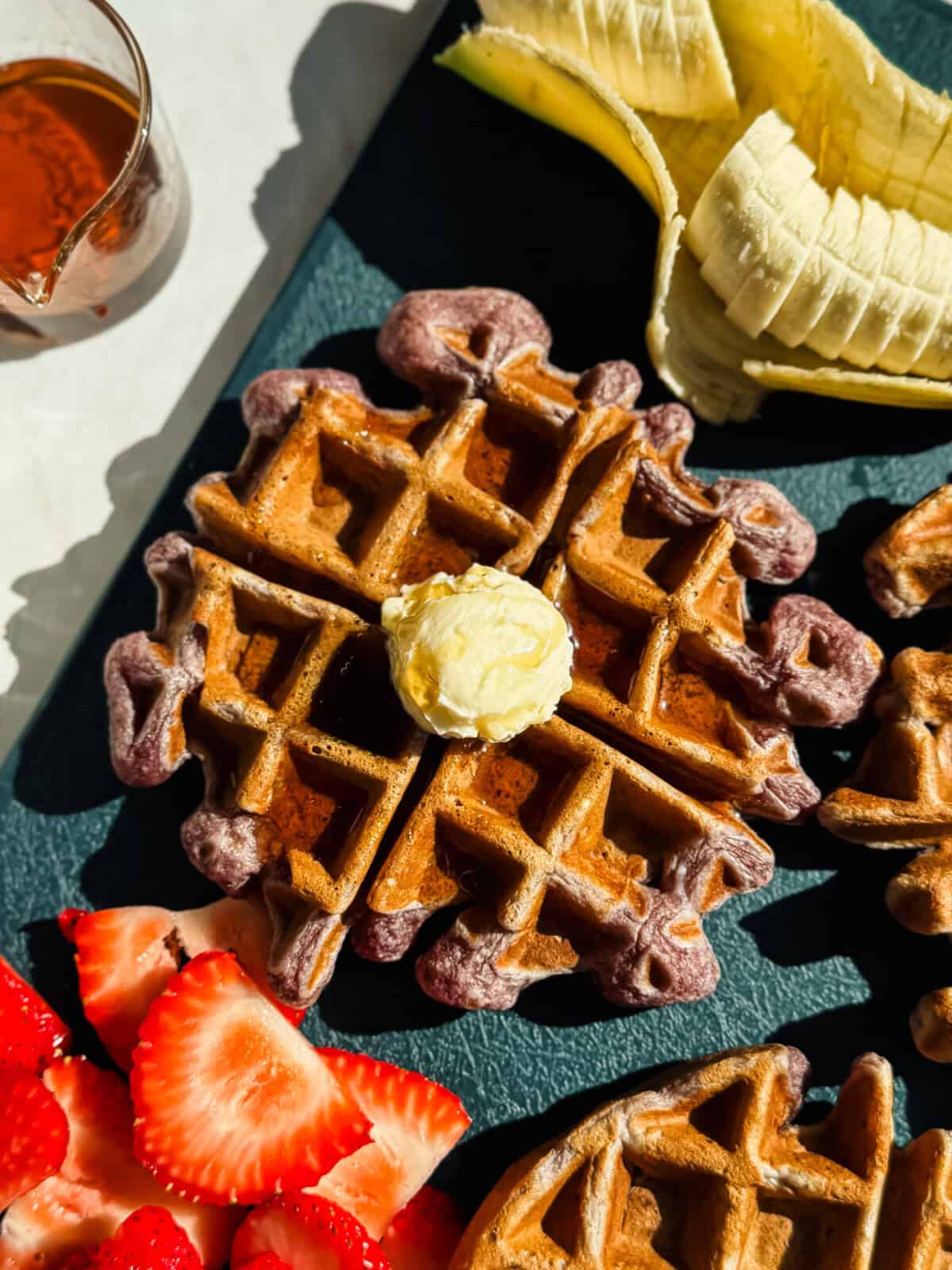 A waffle topped with a pat of butter is shown alongside sliced bananas, strawberries, and a small glass of syrup on a dark surface.