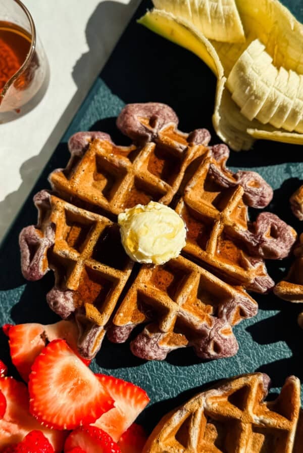 A waffle topped with a pat of butter is shown alongside sliced bananas, strawberries, and a small glass of syrup on a dark surface.
