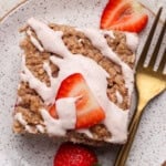 A square piece of oatmeal cake topped with pink icing and sliced strawberries, served on a speckled plate with a gold fork.
