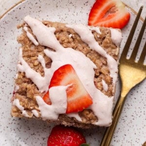 A square piece of oatmeal cake topped with pink icing and sliced strawberries, served on a speckled plate with a gold fork.
