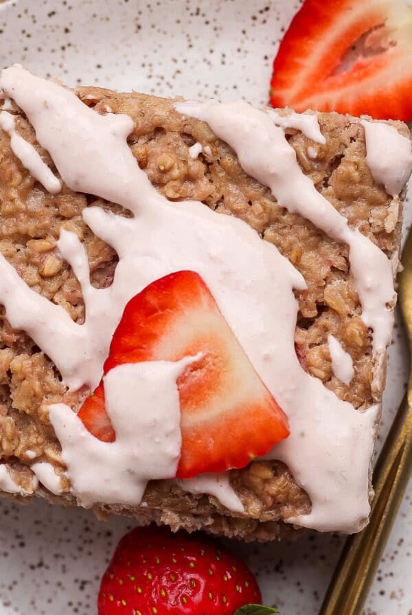 A square piece of oatmeal cake topped with pink icing and sliced strawberries, served on a speckled plate with a gold fork.