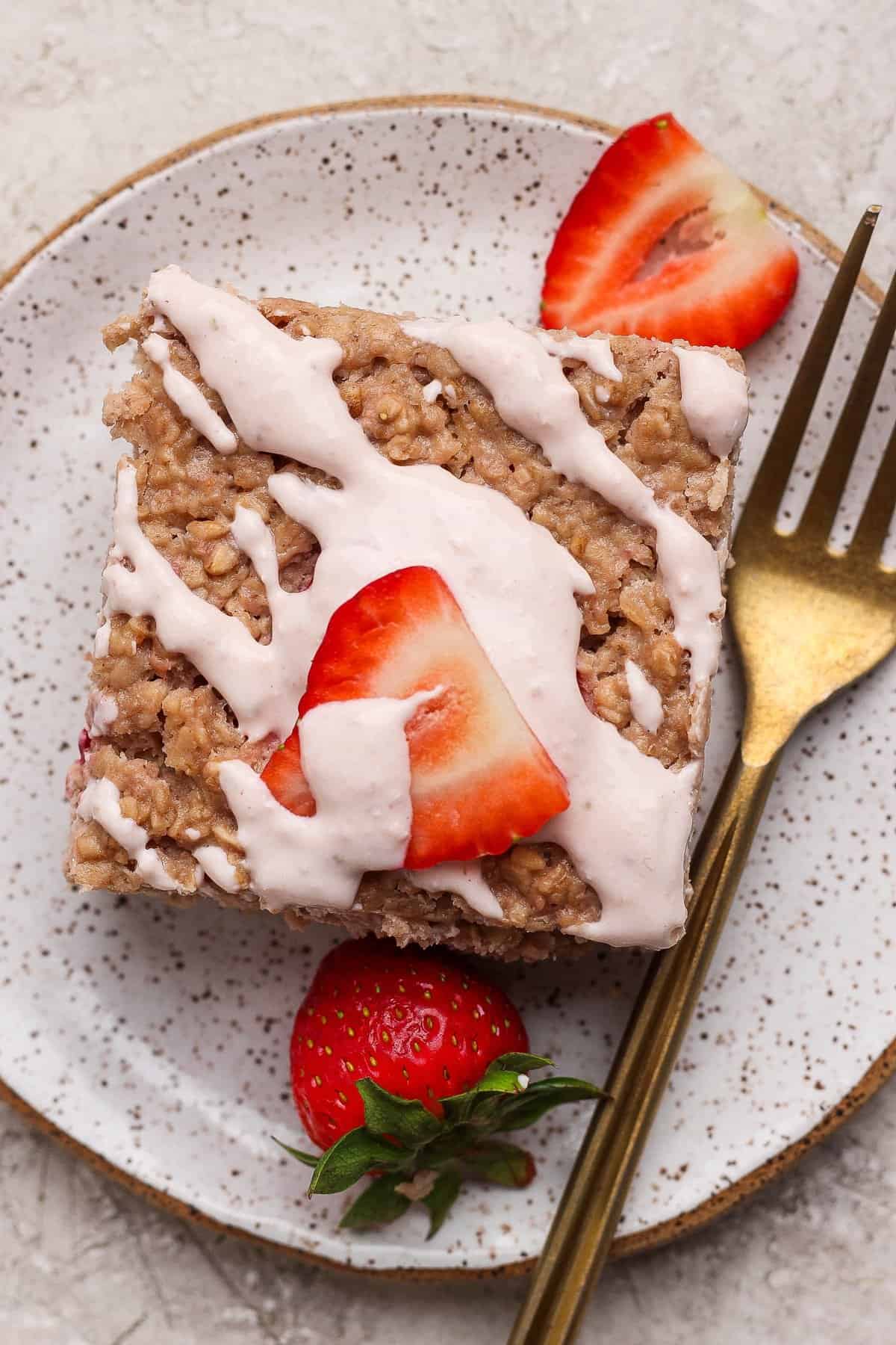 A square piece of oatmeal bake topped with white icing and sliced strawberries on a speckled plate, with a gold fork beside it.
