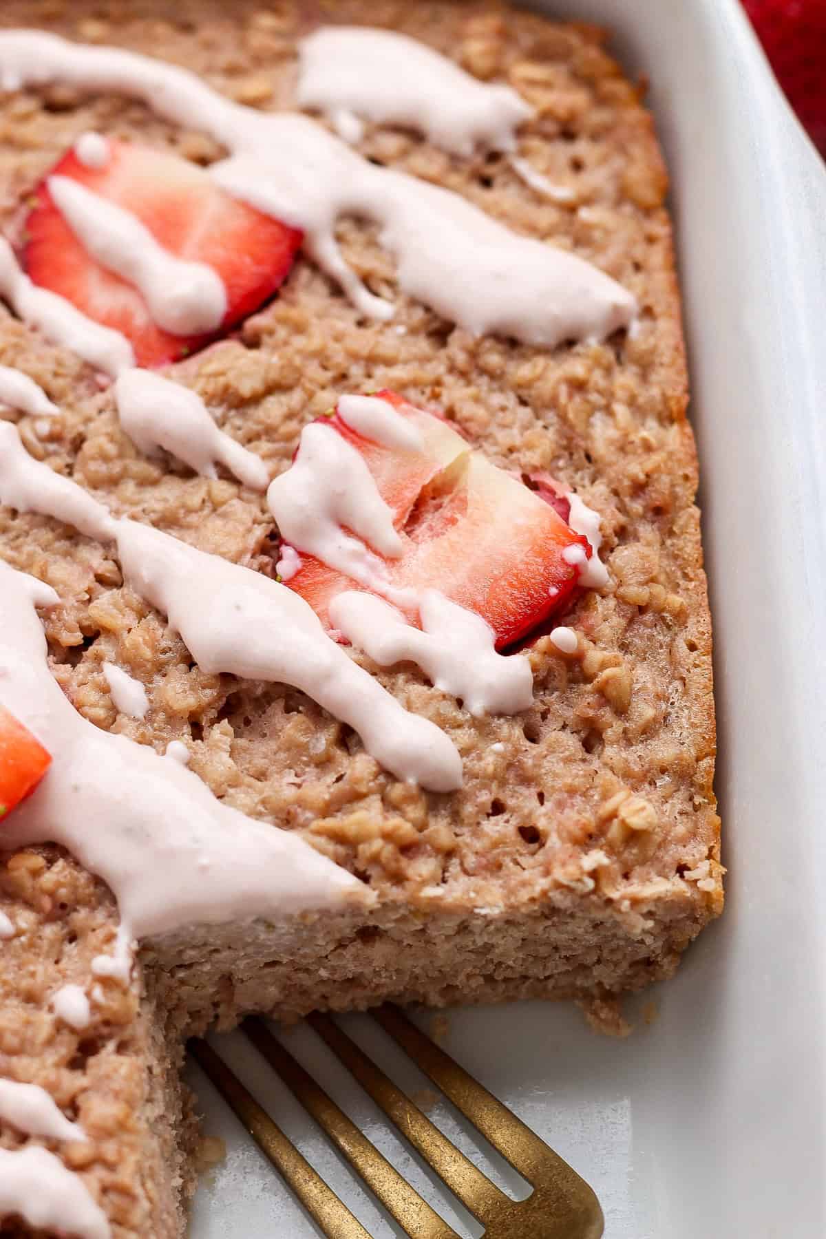 Baked oatmeal topped with sliced strawberries and white icing, shown in a baking dish with a serving removed and a metal spatula visible.
