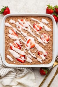 A square pan of baked oatmeal topped with sliced strawberries and white icing drizzle, set on a light surface with whole strawberries, a striped towel, and gold utensils nearby.
