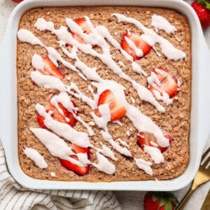 A square pan of baked oatmeal topped with sliced strawberries and white icing drizzle, set on a light surface with whole strawberries, a striped towel, and gold utensils nearby.