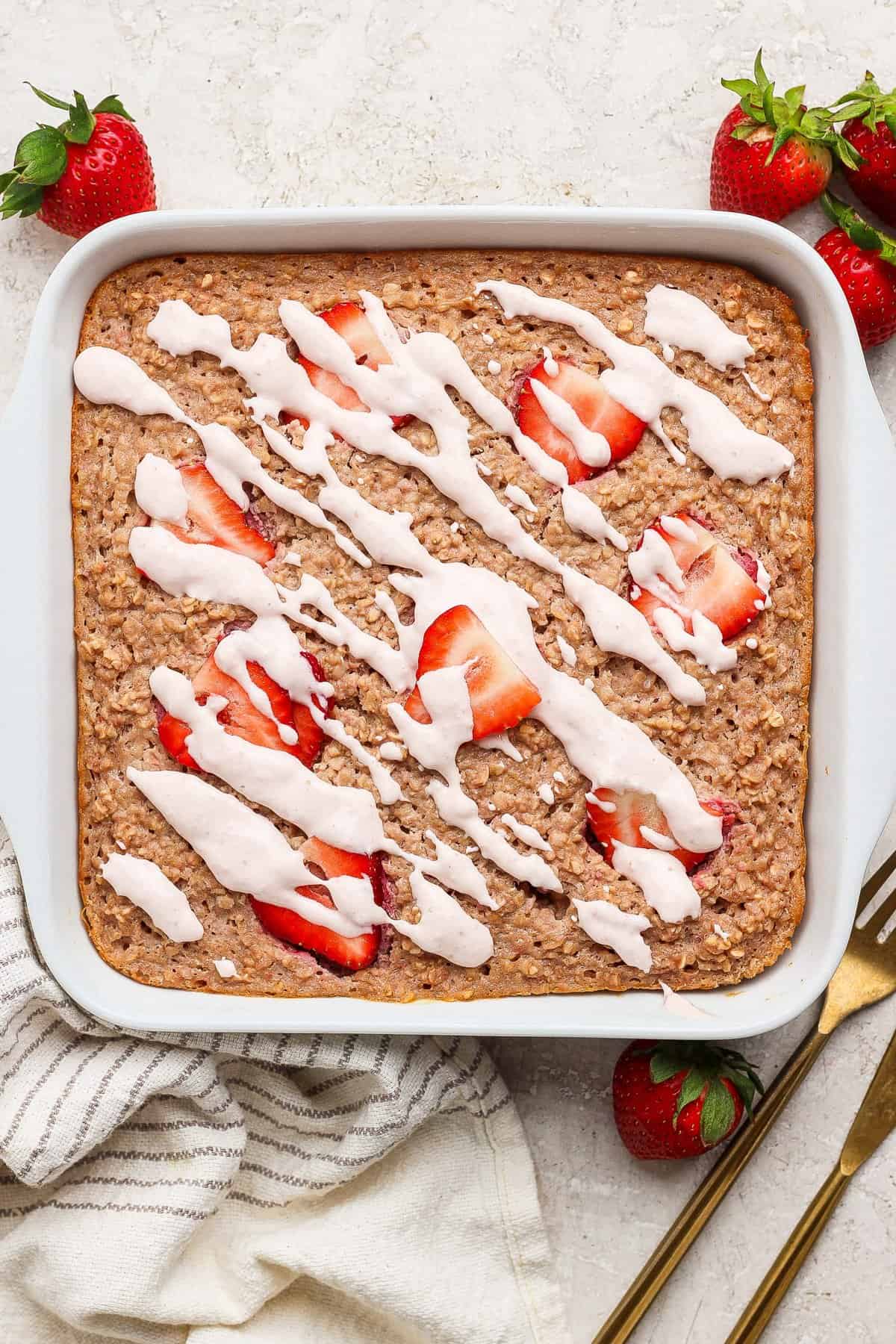 A square pan of baked oatmeal topped with sliced strawberries and white icing drizzle, set on a light surface with whole strawberries, a striped towel, and gold utensils nearby.