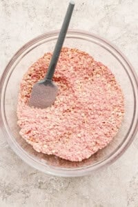A glass bowl filled with pink-tinted oats and a gray spatula resting inside, placed on a light textured surface.
