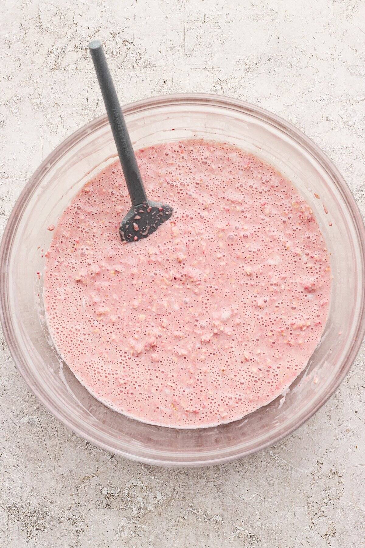 A glass bowl filled with a pink, bubbly batter mixture and a black spatula, placed on a light textured surface.