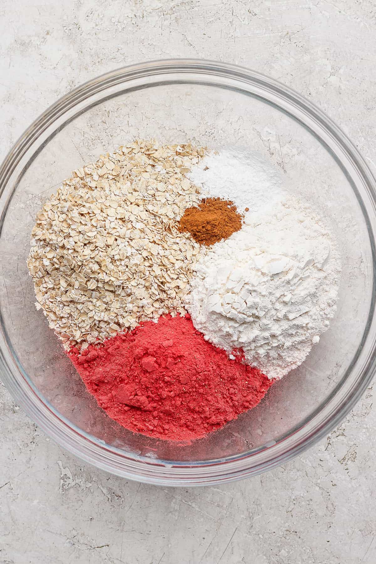 A glass bowl containing oats, white flour, pink powder, cinnamon, baking powder, and baking soda on a light surface.