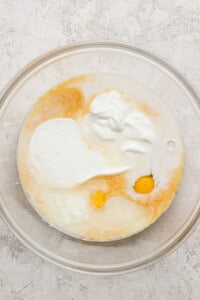 A glass mixing bowl containing an egg, milk, yogurt, and other liquid baking ingredients on a light textured surface.